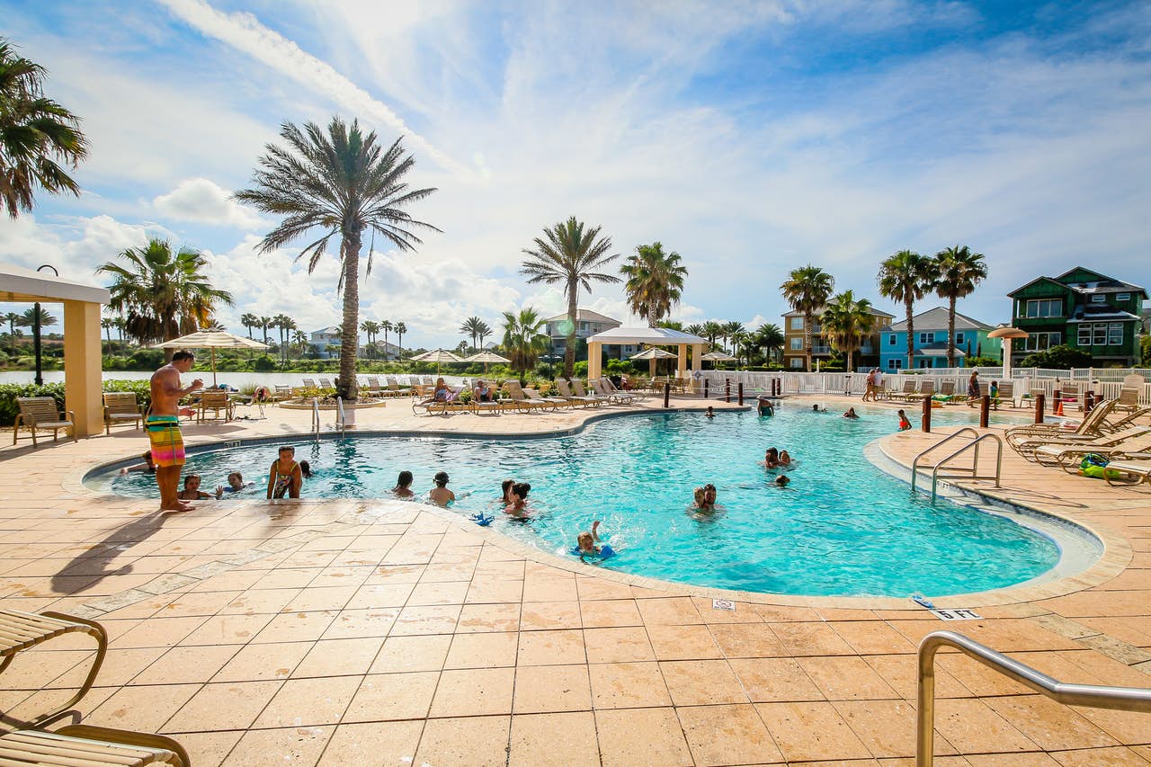 Families playing in a large resort pool near vacation rentals and palm trees on a sunny day.