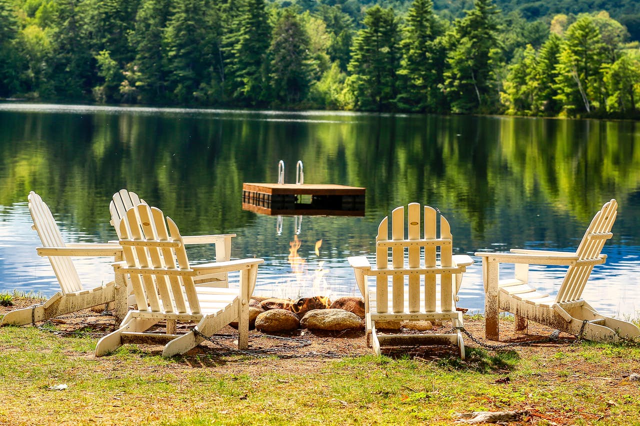 Adirondack chairs surround a fire with a floating dock in the lake beyond