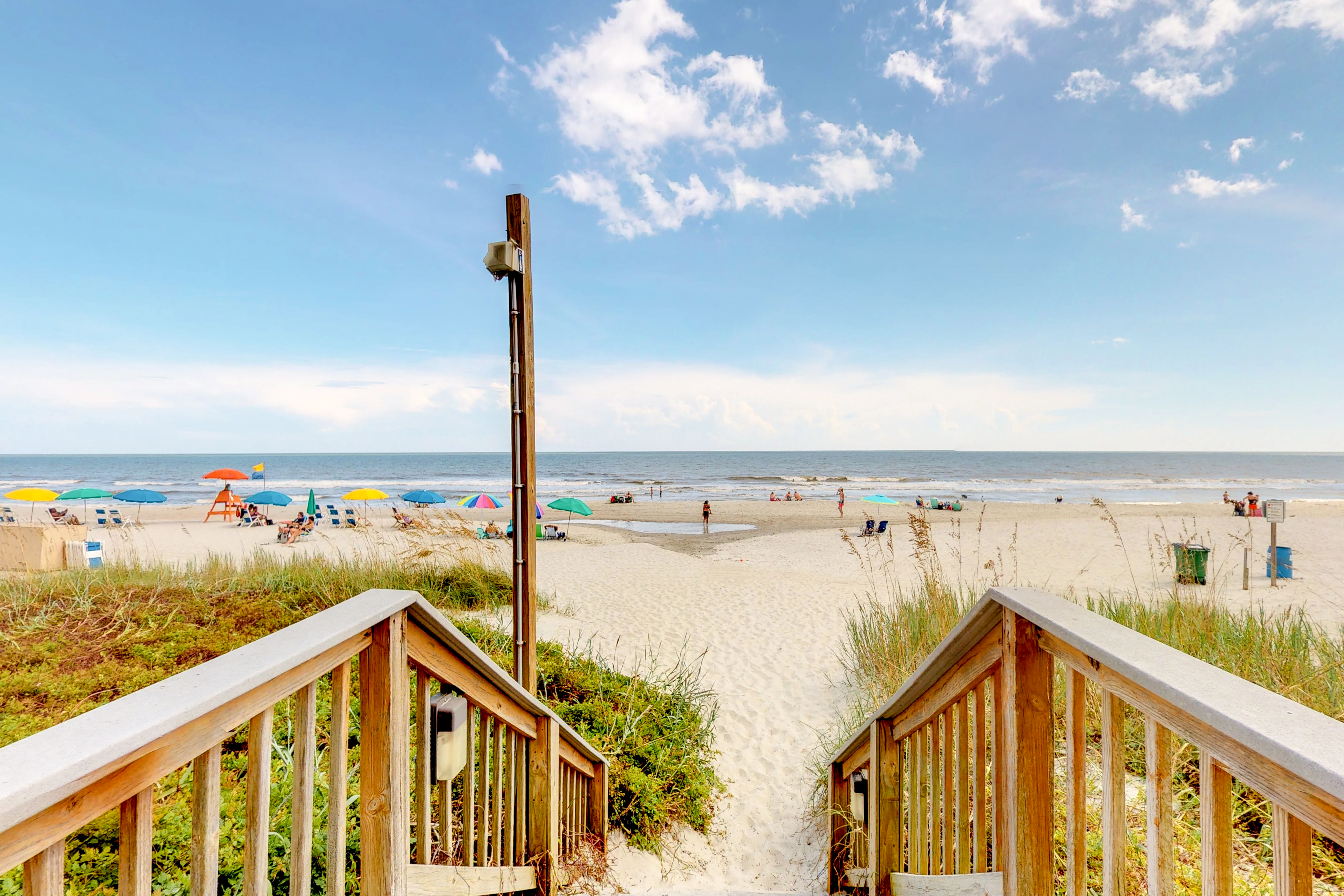 The perspective of a beach from the pathway overlooking the ocean and vacationers under umbrellas.