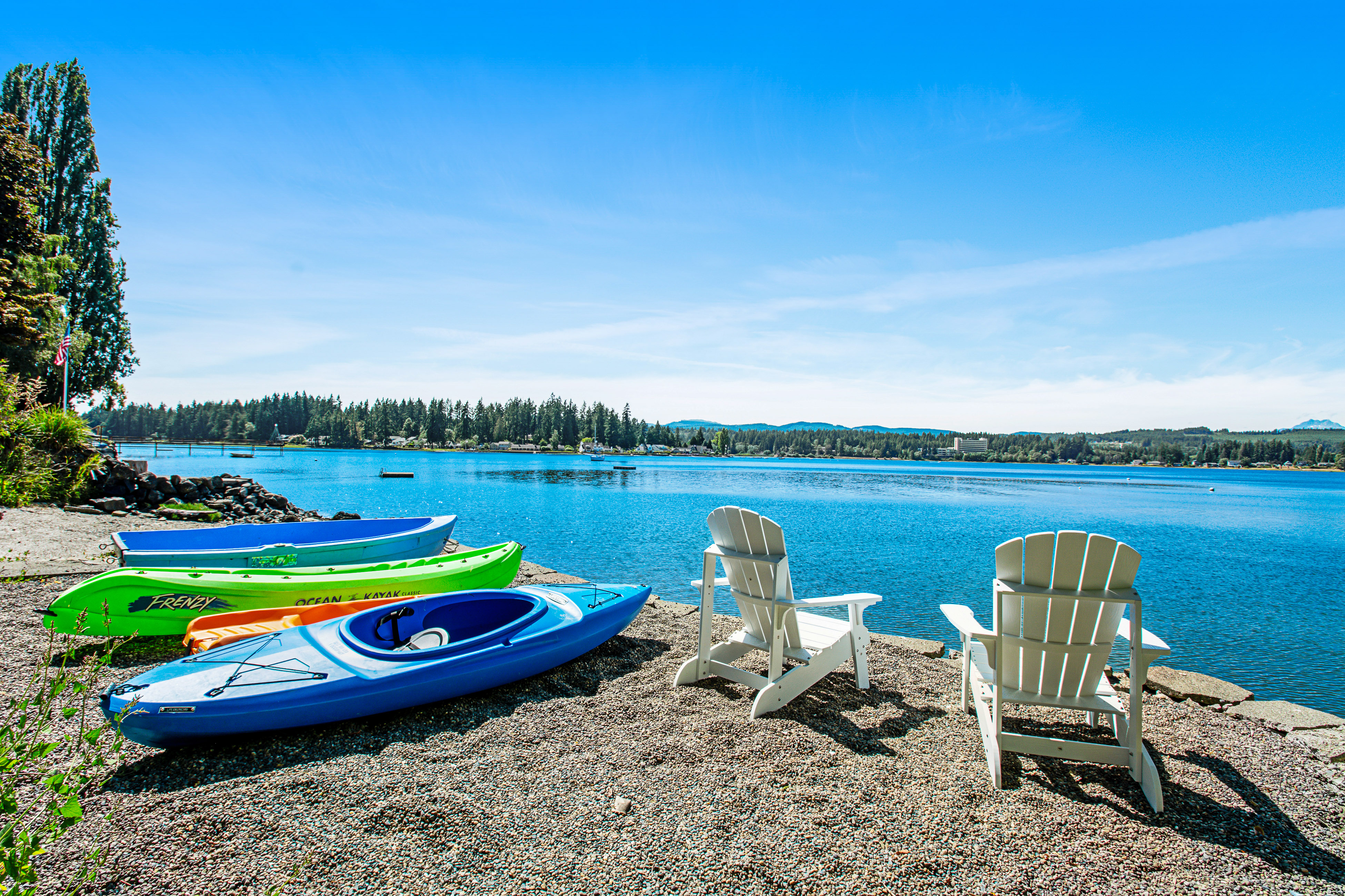colorful kayaks and other boats line a rocky beach next to two white adirondack chairs