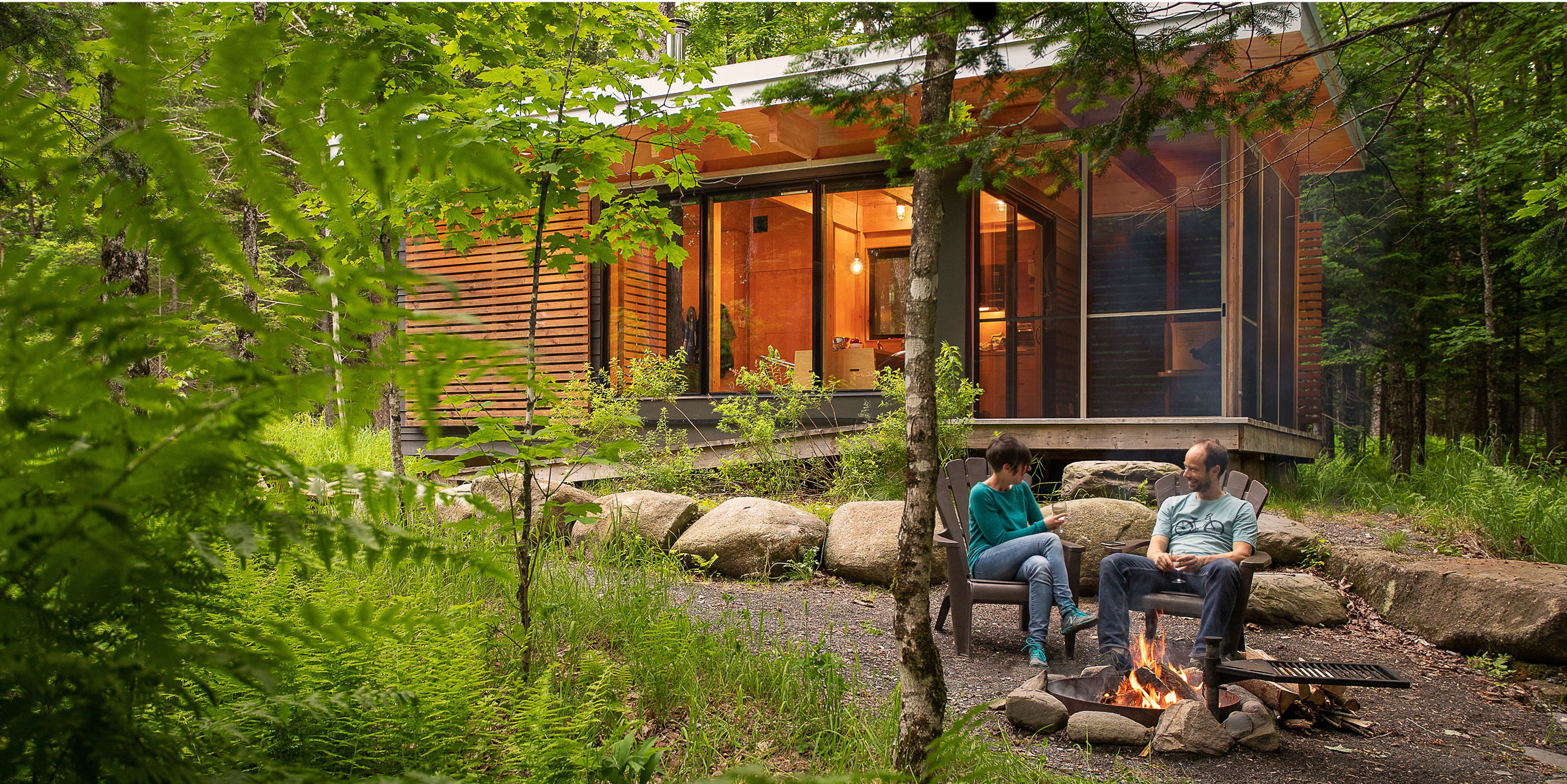 Two people sitting at a camp fire near a cabin.