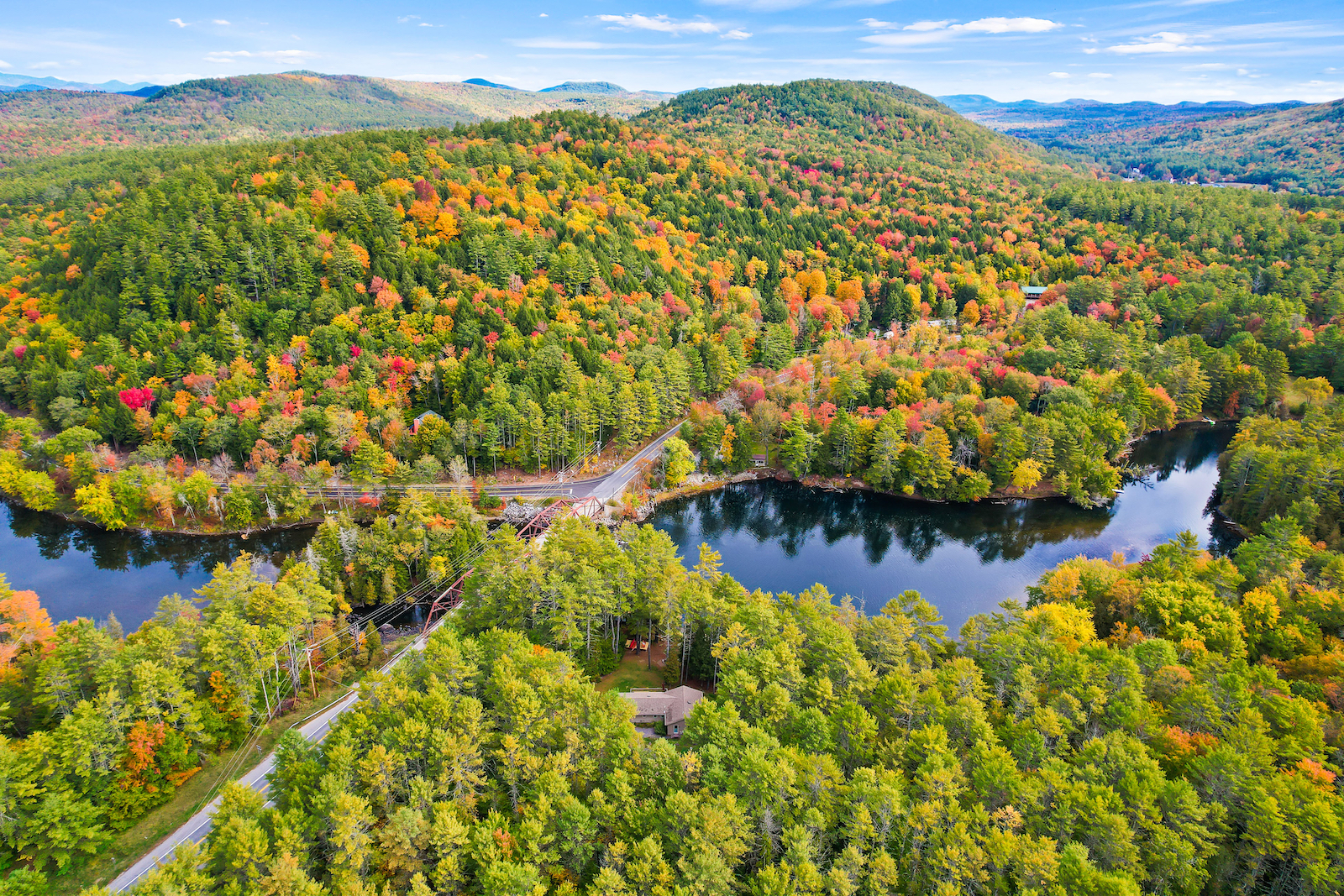 A drone shot of the Adirondacks in the fall overlooking rolling hills, a road, and lakes.