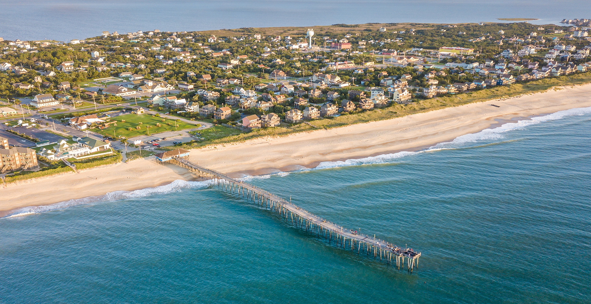 An island with homes and establishments with a pier stretching out to the ocean from the beach.