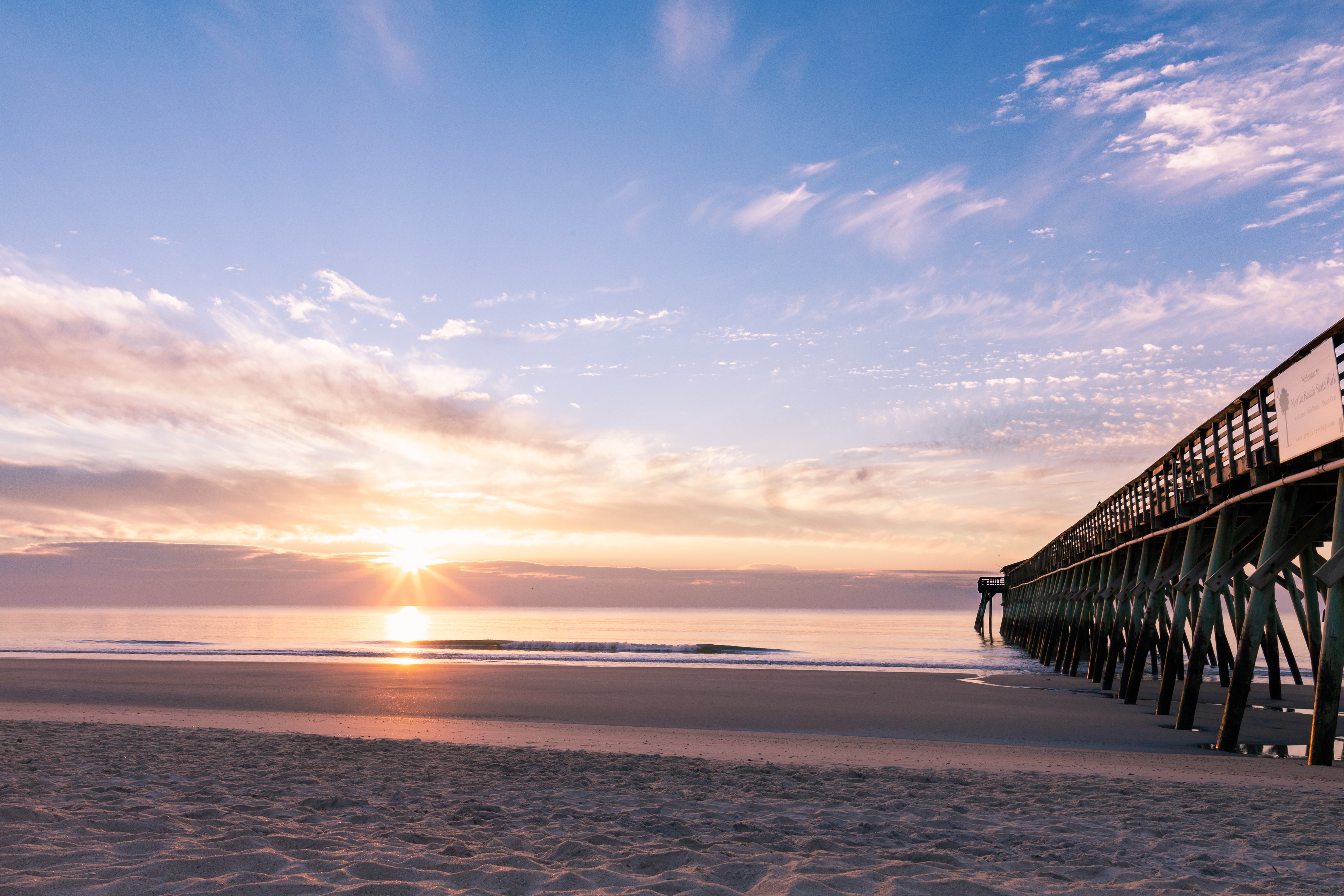 A wooden pier with exposed foundations at a sandy beach during a sunset.