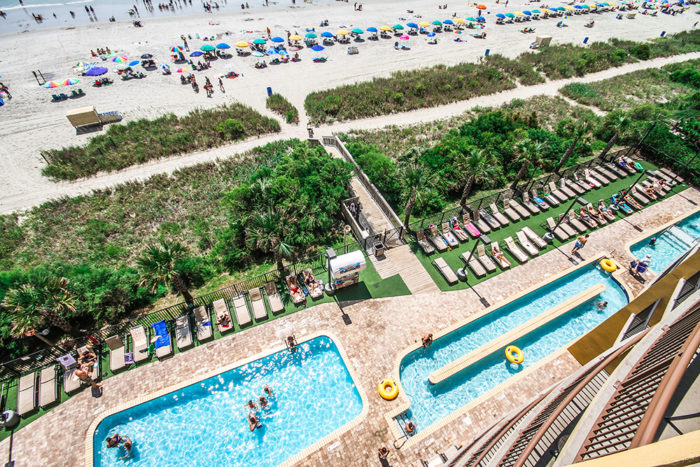 A resort with multiple outdoor pools and rows of pool chairs near a crowded sandy beach.