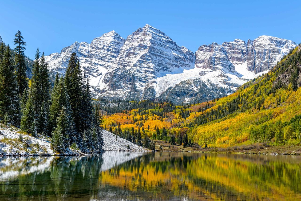 blue skies over a snow capped mountain in aspen, co in front of a lake surrounded by pine trees and fall colors