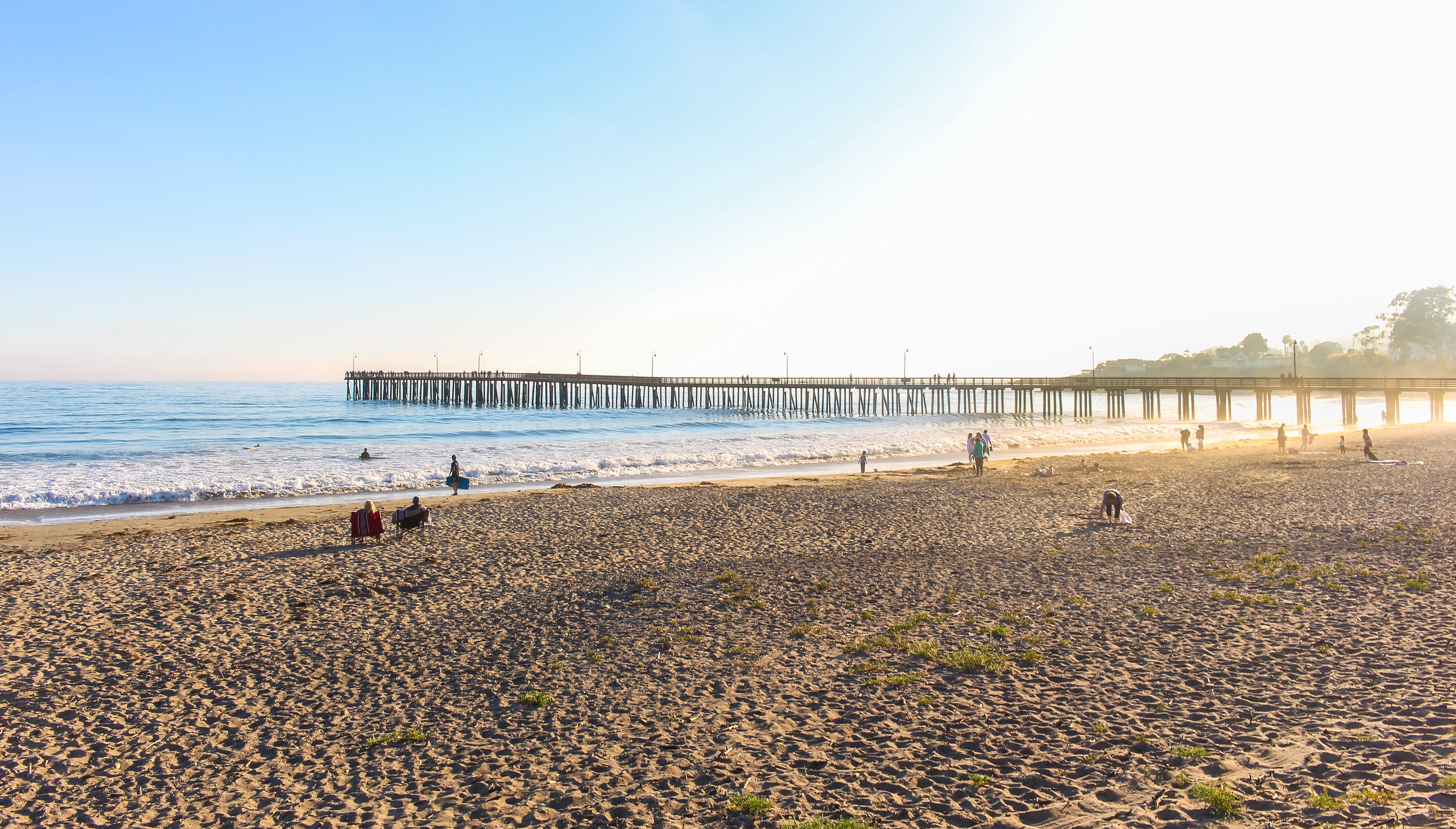 People hanging out on a sandy beach near the shore with a wooden pier out leading out into the ocean.
