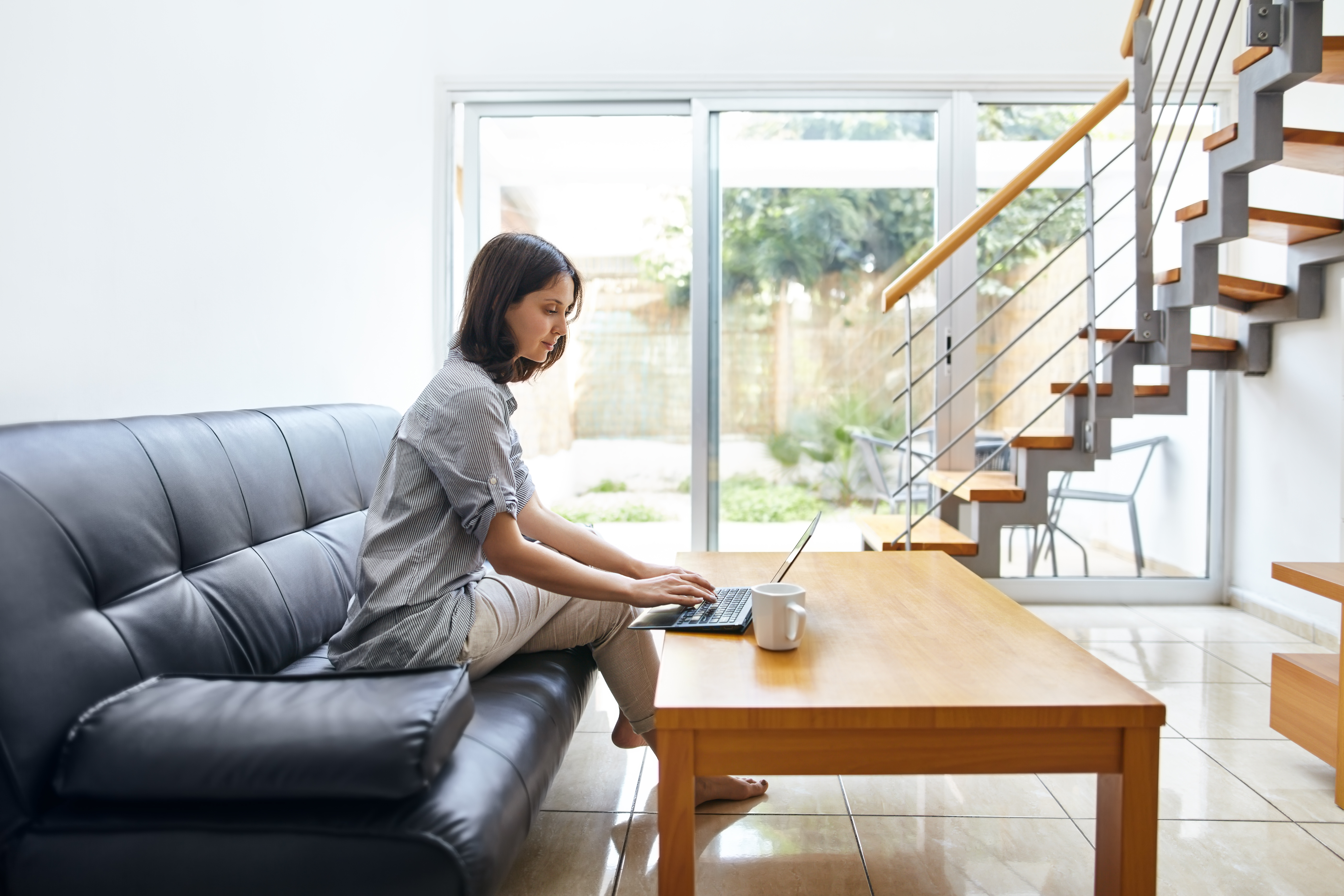 woman sits on a black leather couch while typing on her laptop