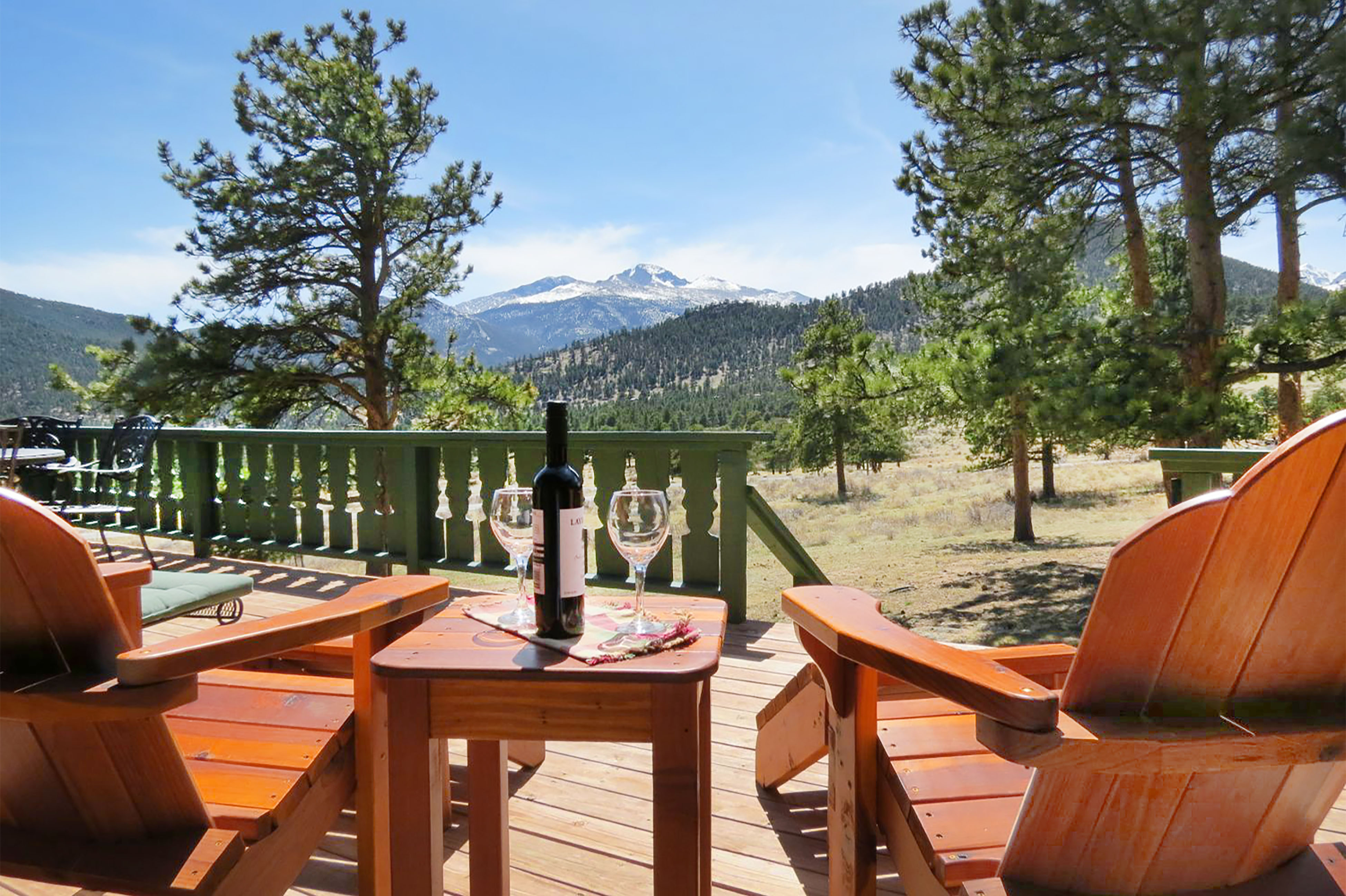 Adirondack chairs on an outdoor deck with a bottle of wine and glasses on a table facing the Rocky Mountains