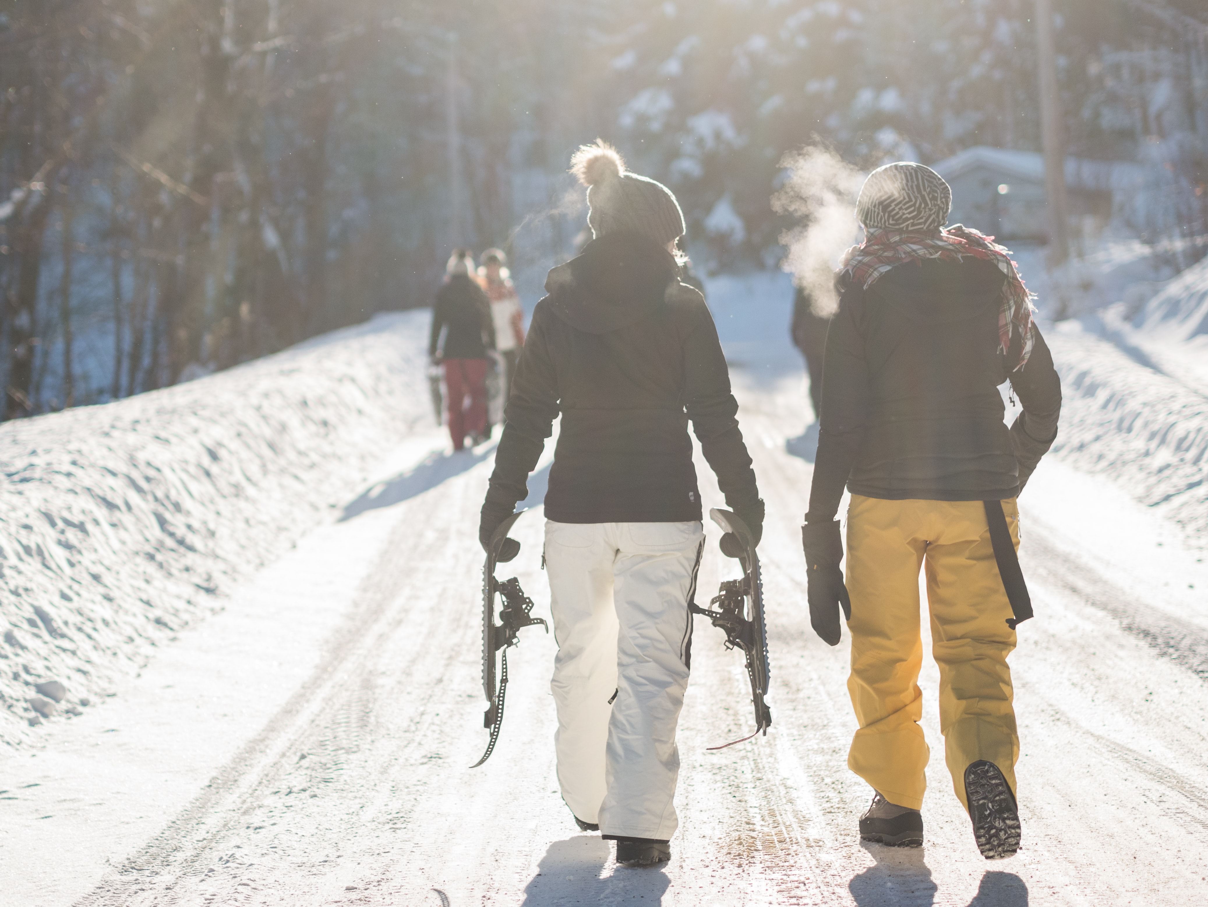 Friends walking up a packed snowy trail carrying snowshoes on a sunny day.
