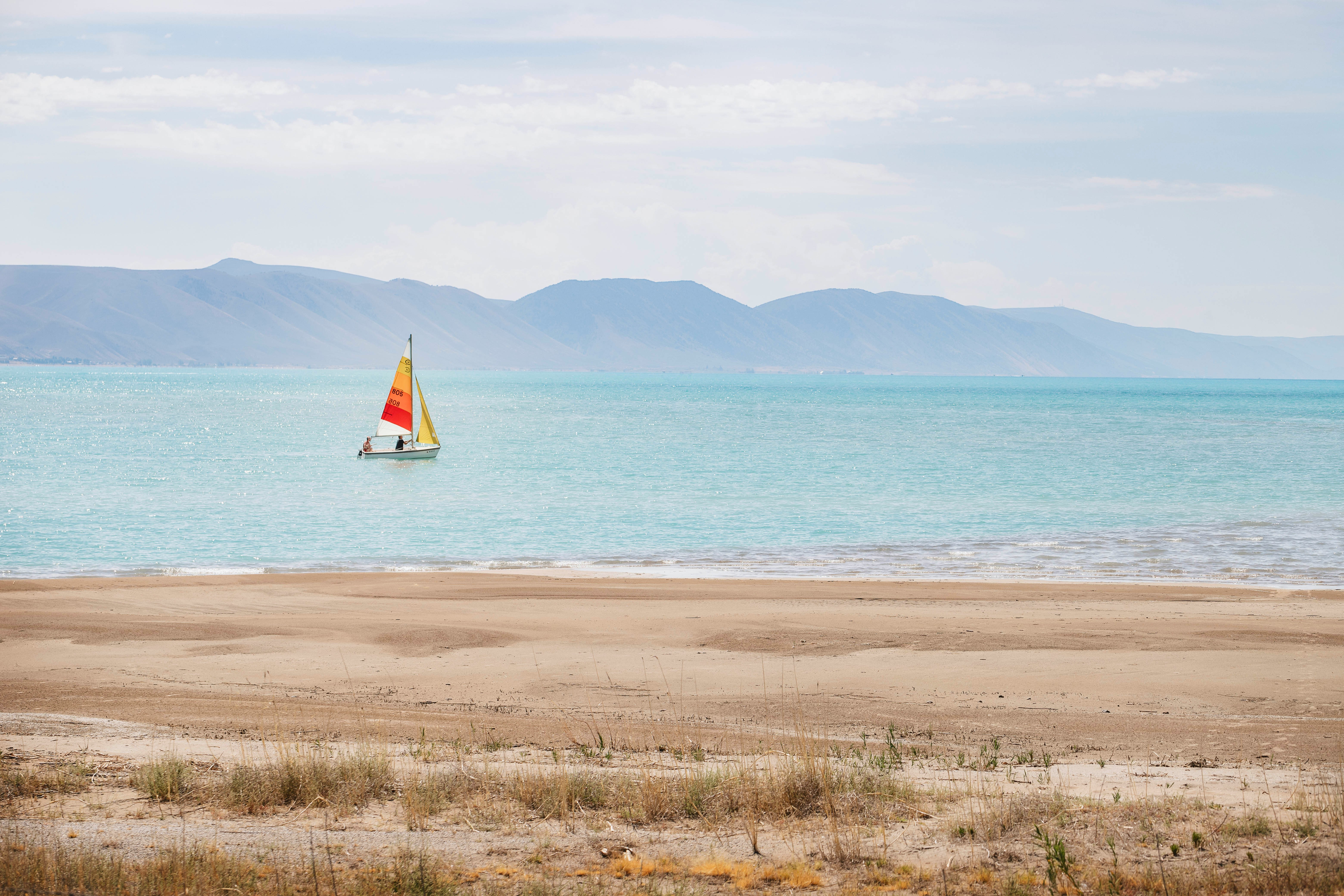 view from a sandy beach of a sailboat with two people on board on bear lake in summer