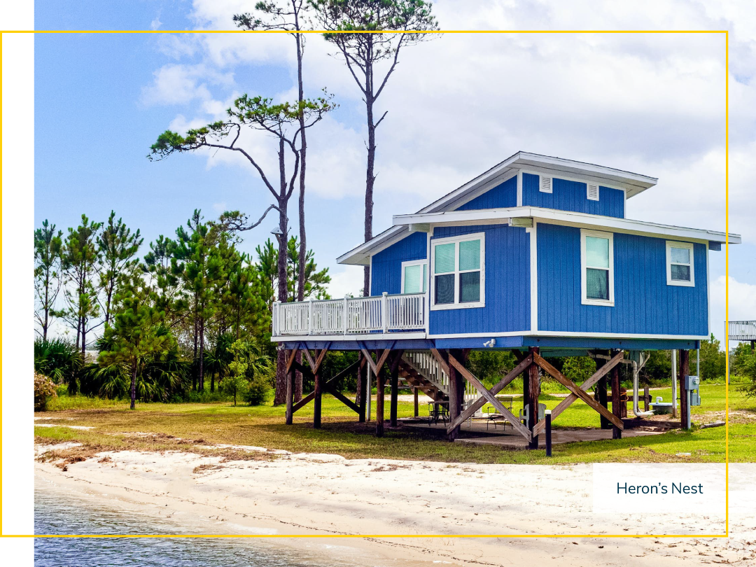 A blue vacation home built on tall wooden foundations near the shore of a beach.